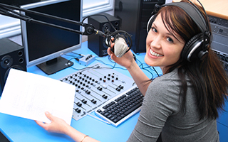 Woman with headphones speaking into a microphone at a radio station booth with audio equipment