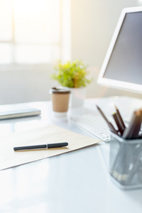 Bright office desk with tablet computer, coffee cup, stationery holder and paper with pen near window with sunlight streaming in
