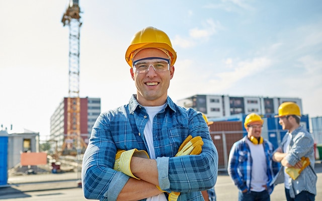 Smiling young man in a plaid shirt and yellow hard hat looking at paper blueprints in front of a construction crane outside