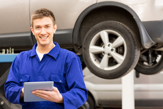 Male automotive technician in blue coveralls smiling and holding a clipboard while standing in front of a hoisted minivan