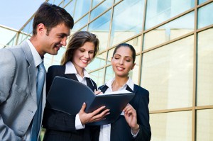 Three business professionals, two women and one man, reviewing documents in front of a modern glass building.