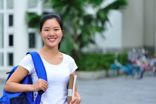 Young female student with backpack holding books, smiling outside college campus