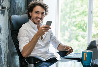 Smiling young man with curly hair using smartphone and laptop at office desk