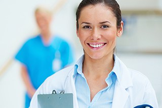 Female healthcare professional with clipboard standing in hospital hallway, male colleague in background.