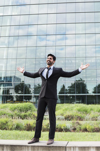 Smiling young man wearing a dark suit and standing in front of a glass building with his arms stretched out to the sides