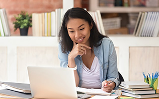 Young woman sitting at a desk in a room full of books and writing notes while smiling at an open laptop