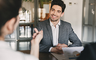 Young man wearing a white shirt and grey suit sitting in an office and smiling at a woman across a table