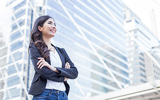 Smiling young woman looking upwards with her arms crossed while standing outside a tall glass building