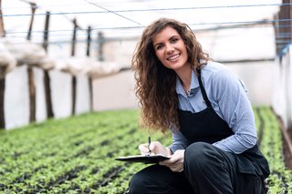 Woman with curly hair smiling and holding clipboard in a greenhouse with young plants.