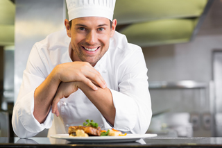 Smiling Chef in white uniform presenting a dish in a professional kitchen.