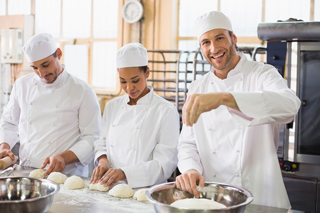 Three chefs in a commercial kitchen preparing dough and smiling, with one gesturing toward the camera
