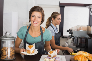 Young woman in apron serving muffins at a bakery counter with another employee preparing coffee in the background