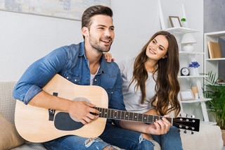 Man playing an acoustic guitar while a woman watches appreciatively in a cozy living room setting.