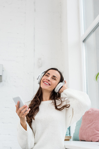 Woman in white sweater enjoying music with headphones and holding smartphone in a bright room.