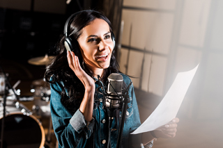 Female singer recording vocals in studio with microphone and headphones, holding sheet of lyrics, smiling and engaged in performance.