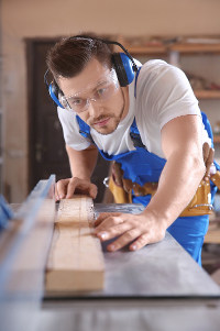 Carpenter using a circular saw to cut a wooden plank in a workshop, wearing safety glasses and hearing protection