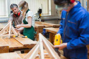 Carpentry workshop with a man instructing a young woman on building a wooden frame while another man works with a power tool in the background.