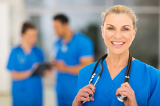 Nurse with stethoscope in blue scrubs, standing in hospital corridor with colleagues in background.