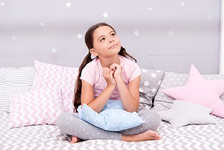 Young girl sitting on bed with star-patterned pillows looking upward with a hopeful expression