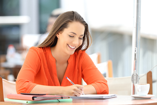 Smiling young woman in a reddish-orange blouse writing in a notebook while sitting at a wooden table with a cup of coffee