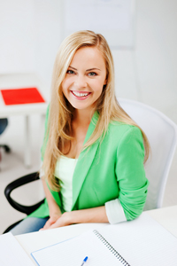Smiling young woman wearing a green semiformal blazer while sitting in a white chair at a desk with an open notebook
