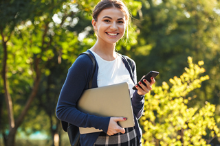 Smiling young female college student carrying a laptop and smartphone outside with trees in the background