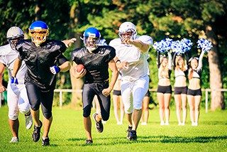 Youth football players in helmets running with the ball during a game with cheerleaders in the background on a sunny day