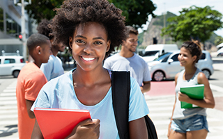Smiling young black woman holding a red notebook crossing the street with other students in the background.