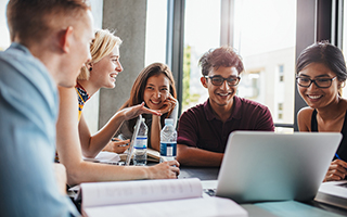 Group of five college students smiling and collaborating on a project around a laptop in a bright classroom.