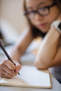 Young woman with glasses writing in a notebook at a desk, focused and thoughtful.