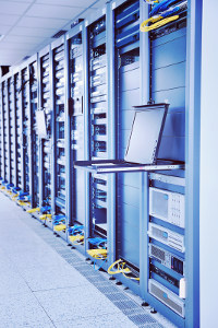 Open laptop on sliding shelf in server room with racks of network equipment and cabling.