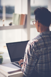 Young man in glasses and plaid shirt working on laptop at desk near window in bright room.