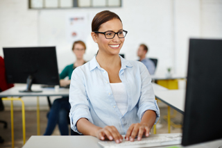 Woman with glasses working at computer in office setting with colleagues in background.