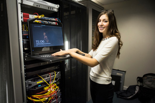 Young woman using computer in server room, monitoring network status on screen, surrounded by equipment and cables.