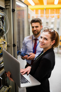 Two IT professionals working in a server room, woman using a laptop.