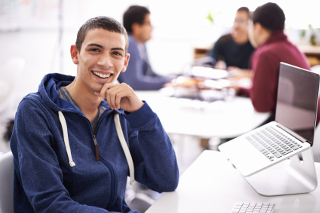 Smiling young man with a laptop at a busy office table, wearing a blue hoodie