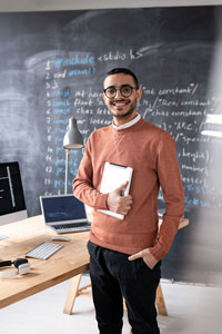 Male teacher with glasses holding a tablet in a classroom with a blackboard in the background.