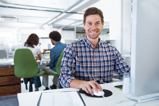 Man in a plaid shirt smiling and using a computer in a well-lit office with two people sitting at another computer behind him