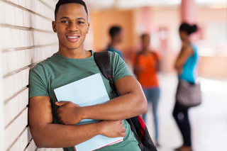 Young man smiling holding a laptop with students in background at a university corridor