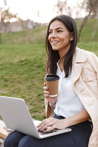Smiling woman sitting on park bench with laptop and holding a coffee cup