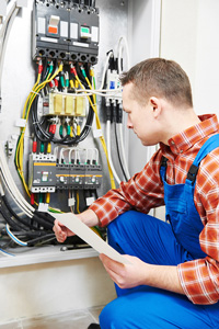 Electrician in checkered shirt and blue overalls examining and maintaining a complex electrical panel indoors
