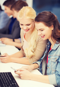 Two young women in a classroom setting. One with a smartphone and the other on a laptop, smiling and engaged in their tasks.