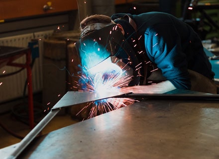 Welder in protective gear performing welding work with bright blue sparks flying on metal workshop table.