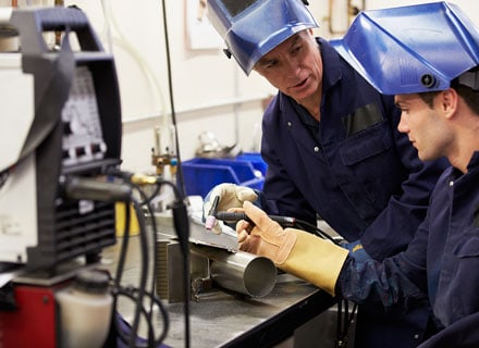 Two welders in blue uniforms and helmets examining a metal part in a workshop