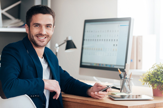 Businessman in a blue jacket sitting at a desk with a computer monitor, smartphone, and desk accessories in an office setting.