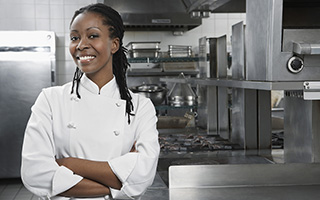 Smiling female chef in a white uniform standing in a commercial kitchen with her arms crossed in front of her