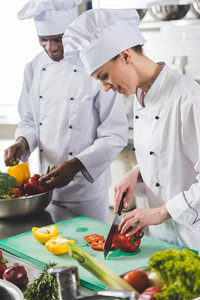 Two professional chefs in a kitchen, a woman slicing red bell peppers and a man slicing vegetables in the background, both wearing white chef uniforms and hats.