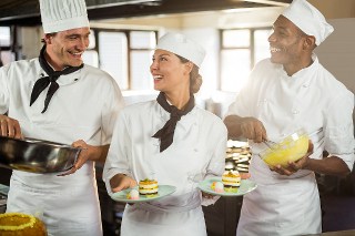 Three professional chefs in a kitchen, laughing and preparing desserts, with one holding a frying pan and another presenting a plate of colorful layered cakes.