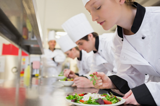 Row of three young adults in chef uniforms intently finishing plated dishes of food while a chef observes in the background
