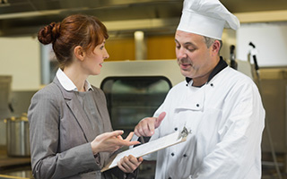 Chef in white uniform discussing menu with female manager in restaurant kitchen.